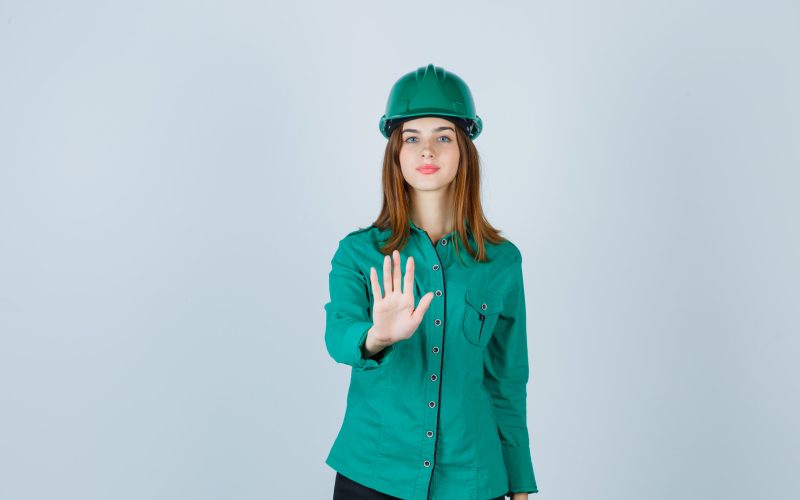 young lady in shirt, pants, helmet showing stop gesture and looking serious , front view.
